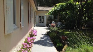 Un sentiero stretto, costeggiato da fiori colorati e vegetazione, conduce a un edificio con tetto in tegole e persiane bianche.