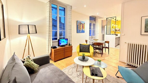 A modern living room with a sofa, coffee table, and TV, a dining area with chairs, and a kitchen visible through an open door.