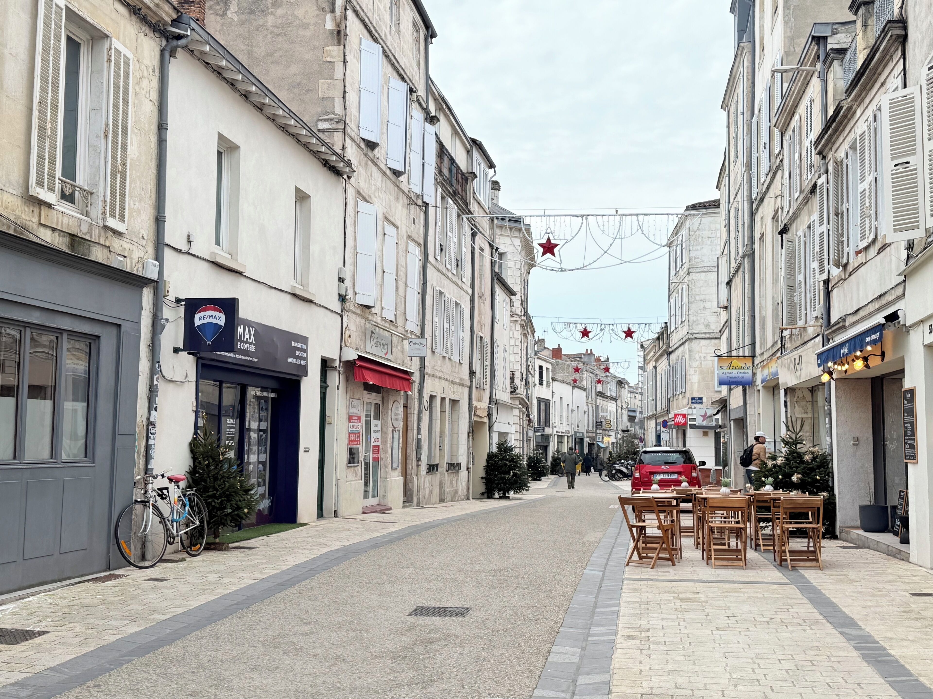 Une rue pavée bordée de bâtiments historiques, un café avec des tables en terrasse et une enseigne RE/MAX.