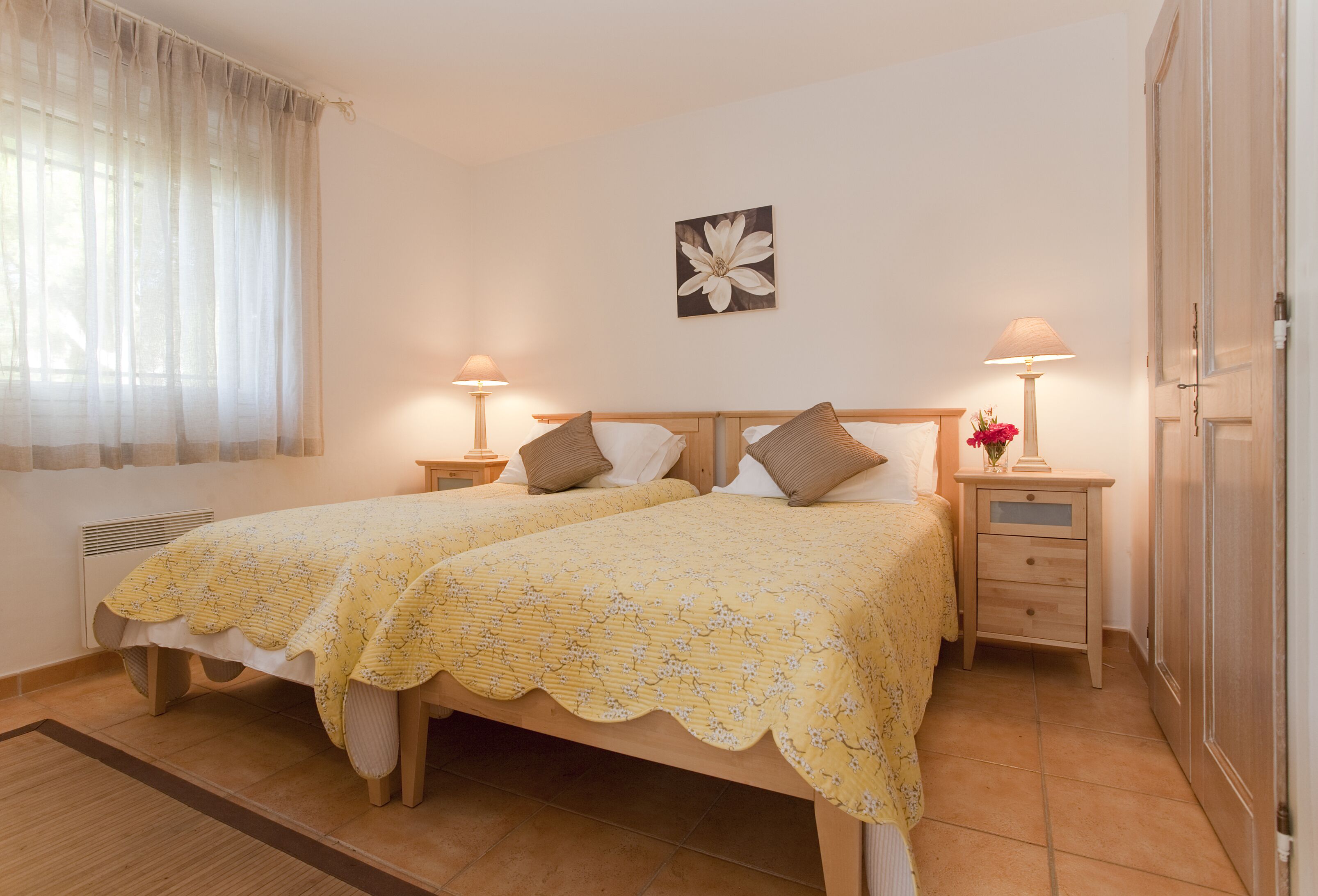 Un lit double avec une couvre-lit à motifs floraux, deux tables de chevet avec des lampes, un tableau représentant des fleurs accroché au mur et un radiateur.