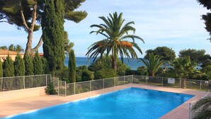 A rectangular swimming pool with a white metal fence, surrounded by various trees and a view of the sea. - 4 bed sleeps 8  beach front villa with Shared Pool and Private Garden. (Roquebrune-Sur-Argens)