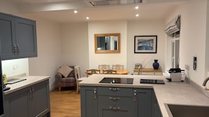A compact kitchen with grey cabinets, a dining area with a wooden table and chairs, a mirror, and a framed picture on the wall. - Hill Rise - Cottage In beautiful Trevone Bay, Near Padstow, North Cornwall (Trevone, Padstow)