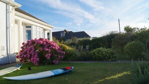 A white building with a large window and a door, a bush with pink flowers, and a surfboard on the grass. - Hill Rise Cottage in beautiful Trevone Bay, near Padstow, North Cornwall (Trevone, Padstow)