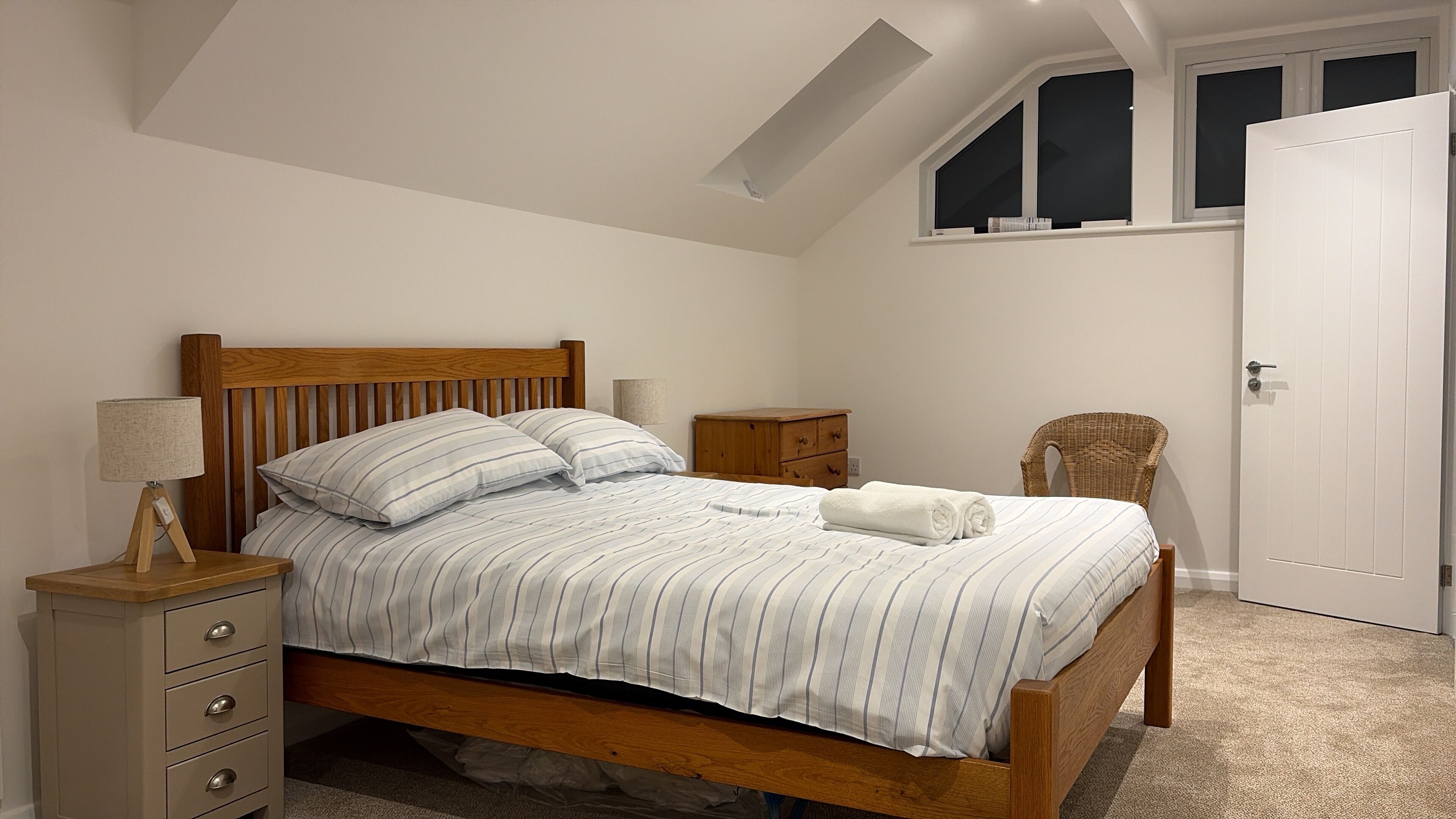 Une chambre à coucher avec un lit en bois, une table de chevet avec une lampe, une petite commode en bois et une chaise en osier.