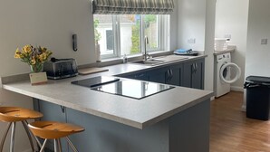 A modern kitchen with a central island, bar stools, a built-in oven, and a window with a striped valance. - Hill Rise Cottage in beautiful Trevone Bay, near Padstow, North Cornwall (Trevone, Padstow)
