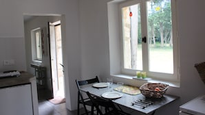 A dining area with a table set for two, a window with a view of trees, and a kitchen area visible in the background. - Shed the heart of Provence (Saint-Rémy-De-Provence)