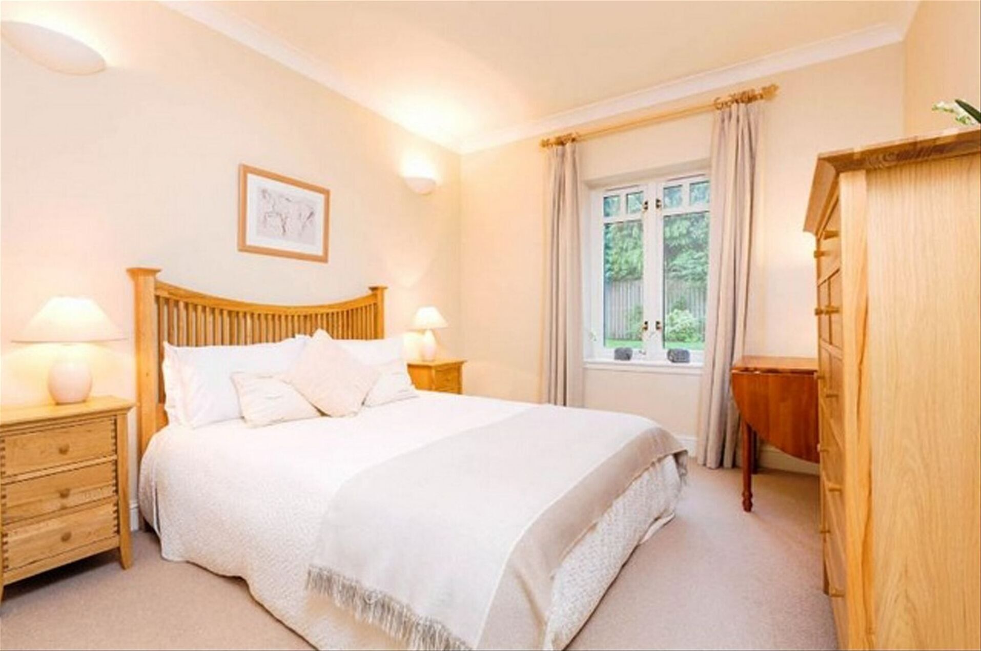 A bedroom with a wooden bed, two bedside tables with lamps, a wooden dresser, a window with curtains, and a framed picture on the wall.