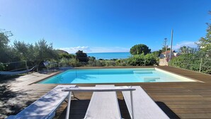 A swimming pool with two sun loungers on a wooden deck, surrounded by greenery and a view of the sea. - Holiday Home Sciacca (San Giorgio)