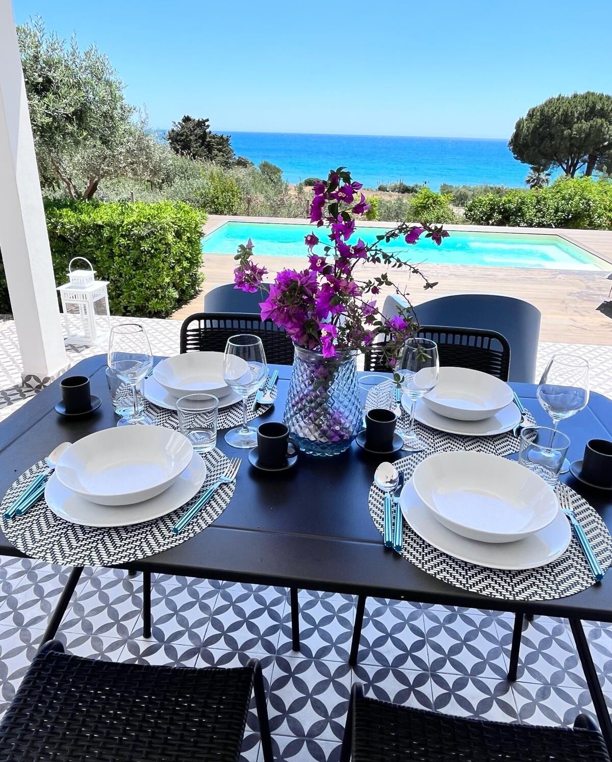 Tavola imbandita per quattro con piatti bianchi, posate nere e un vaso di fiori viola, con vista su una piscina e l'oceano.