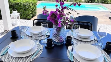 Tavola imbandita per quattro con piatti bianchi, posate nere e un vaso di fiori viola, con vista su una piscina e l'oceano.