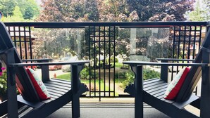 A balcony with two black lounge chairs, each with a red and white pillow. The balcony overlooks a garden with trees and a lawn. - Artisan Suites on Bowen (Bowen Island)