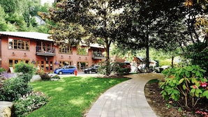 A brick building with a curved driveway, surrounded by lush greenery and parked cars. - Artisan Suites on Bowen (Bowen Island)