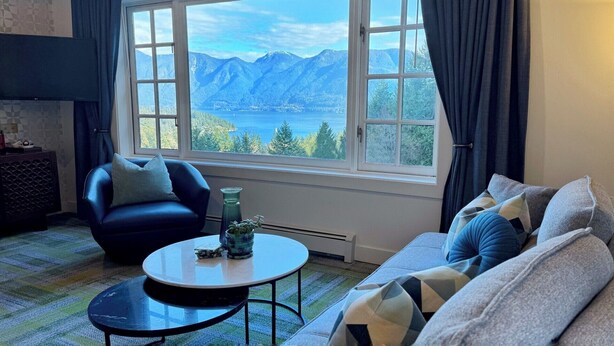 A living room with a large window offering a mountain and lake view, a blue sofa, a round coffee table, and a patterned rug. - Artisan Suites on Bowen (Bowen Island)