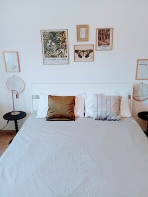 Una cama bien hecha con edredón blanco, almohada marrón y almohadas a rayas. En la pared, encima de la cama, hay impresiones enmarcadas de motivos botánicos.