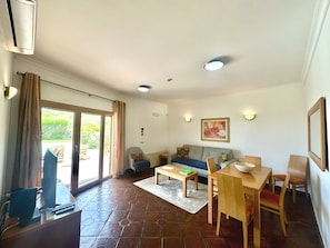 A living room with a sofa, a dining table, and a TV. There is a sliding glass door leading to an outdoor area.