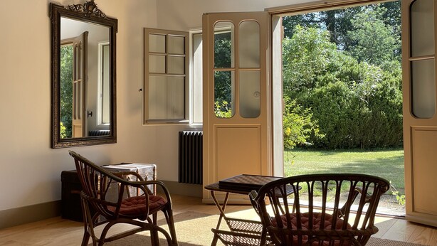 Une pièce spacieuse avec des poutres en bois, un grand miroir, deux fauteuils en osier, une petite table et une vue sur la verdure par des portes ouvertes.