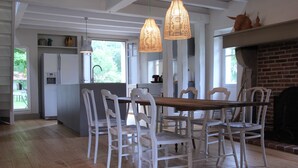 Un coin repas avec une table en bois et des chaises blanches, une cuisine en arrière-plan et une cheminée en briques.