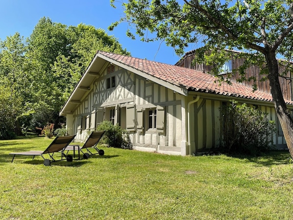 Uma casa de dois andares com telhado inclinado, varanda e gramado com espreguiçadeira.