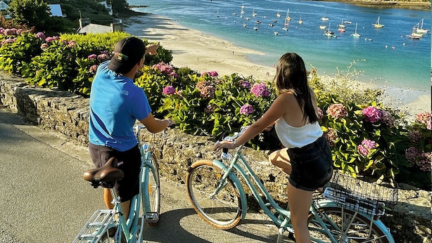 Dos personas en bicicleta mirando una playa con barcos y casas al fondo.