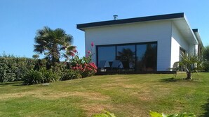 Una casa moderna con un amplio ventanal de vidrio, rodeada de un césped cuidado y plantas tropicales.