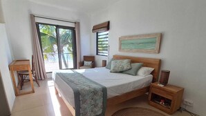A bedroom with a bed, a chair, a nightstand, a window with curtains, and a view of palm trees. - Pointe D Esny: House / Villa - Pointe d''Esny (Pointe d'Esny)