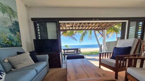 A living room with a sofa, a coffee table, and a TV. There is a view of the beach and palm trees through large windows. - Pointe D Esny: House / Villa - Pointe d''Esny (Pointe d'Esny)