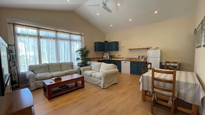 Amplia sala de estar con sofá, una mesa de centro y una mesa de comedor con sillas. Se observa una cocina equipada con microondas, refrigerador y armarios. Un ventilador de techo es visible, y la habitación tiene ventanas grandes con cortinas.