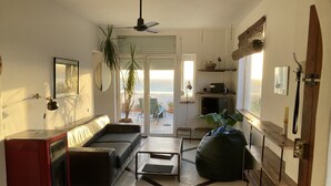 A living room with a sofa, coffee table, and bean bag. There is a TV and a red appliance on the left. A ceiling fan is visible, and the room has a view of the ocean through a door. - SEAFRONT APARTMENT (Tarifa)