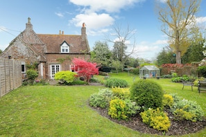 A traditional brick house with a sloped roof, a garden with various plants and a greenhouse, and a bench in the yard.