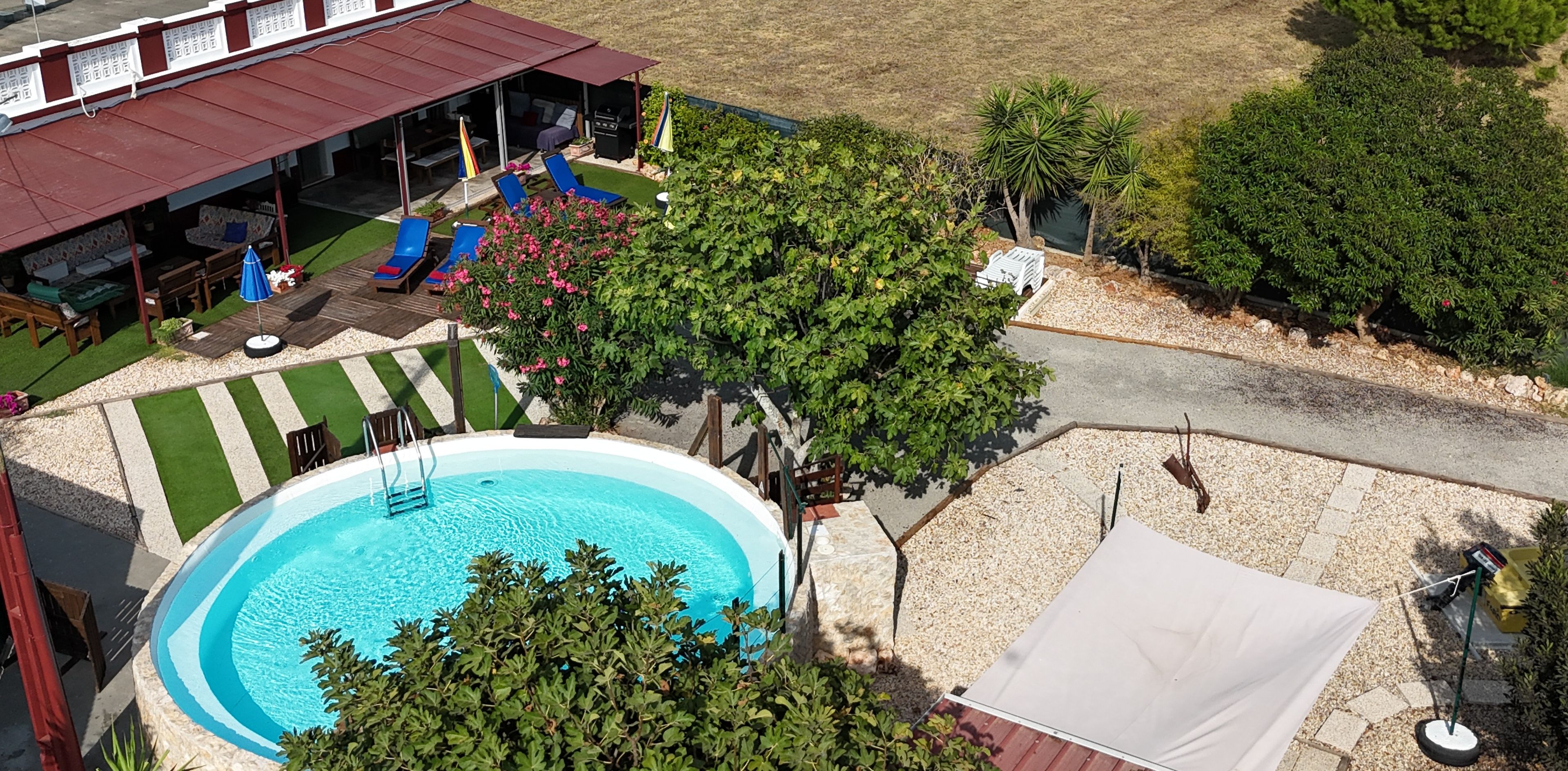Una piscina con zona de solárium, rodeada de vegetación y un área de estar cubierta con sillones.