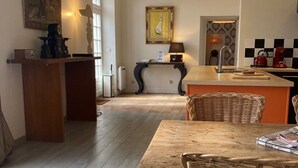 Un salón amplio con una mesa de comedor de madera, sillas de mimbre, una zona de cocina con un hervidor rojo y un cuadro en la pared.