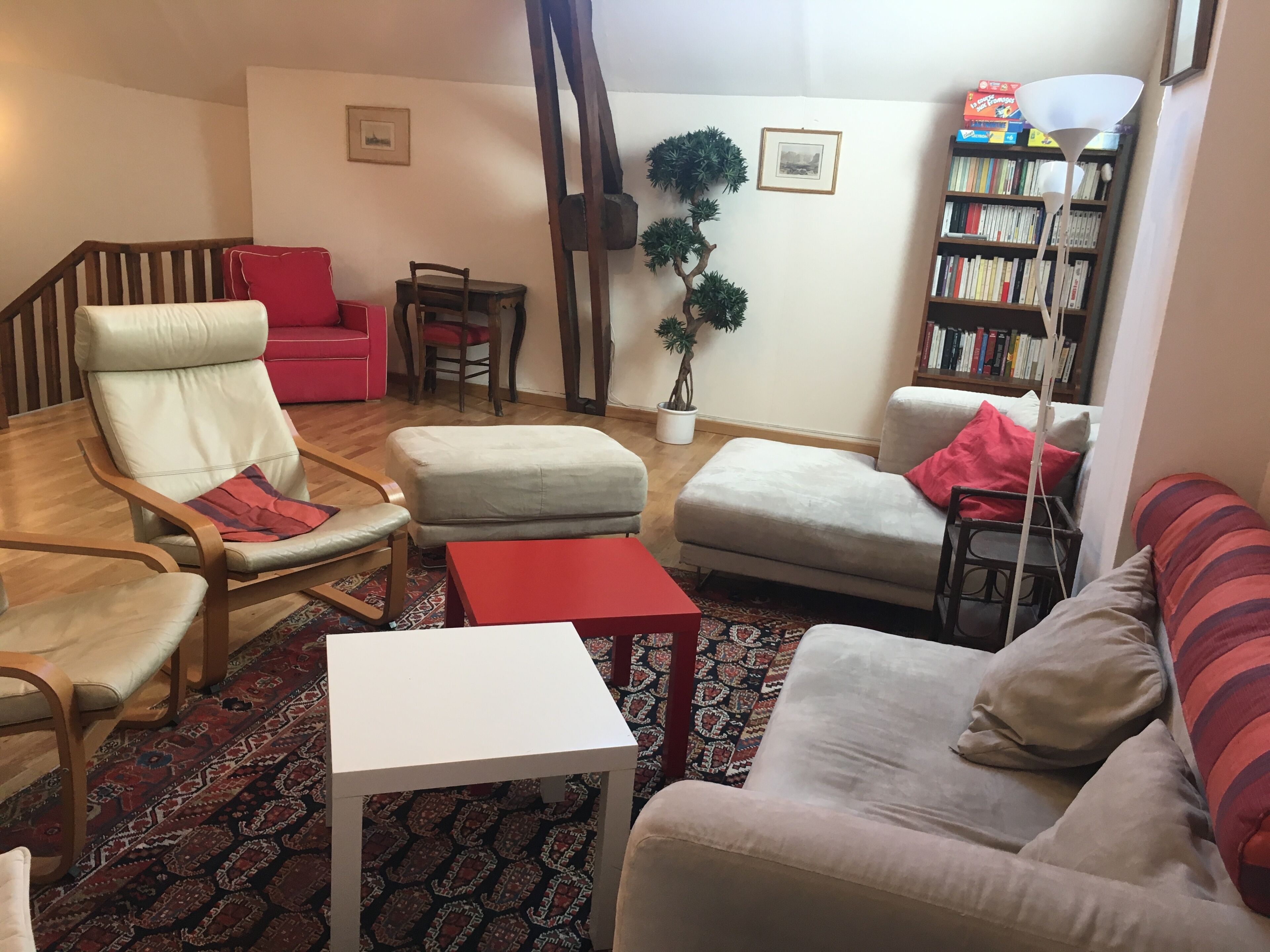 Un salon comprenant un canapé, un fauteuil, un pouf et une petite table. On y trouve également une bibliothèque et une plante en pot.
