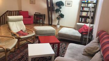 Un salon comprenant un canapé, un fauteuil, un pouf et une petite table. On y trouve également une bibliothèque et une plante en pot.