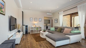 Un salon moderne avec un canapé d’angle, un téléviseur à écran plat, un coin repas avec une table et des chaises, et une cuisine en arrière-plan.