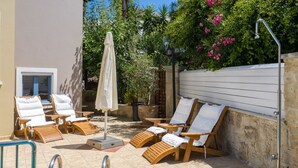 A sunlit outdoor seating area with wooden lounge chairs and a covered umbrella, surrounded by a stone wall and lush greenery. - Villa Kallergi - Nefeli (450 m2) House for 6, west-central Crete (Rethymnon)