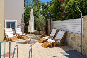 A sunlit outdoor seating area with wooden lounge chairs and a covered umbrella, surrounded by a stone wall and lush greenery.