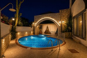 An outdoor swimming pool with a stone wall and archway, surrounded by plants and evening lighting.
