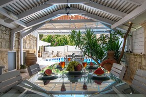 An outdoor dining area with a glass table, white benches, and a view of a pool and greenery. - Villa Kallergi - Nefeli (450 m2) House for 6, west-central Crete (Rethymnon)