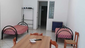 Quarto com mesa de madeira, quatro cadeiras, duas camas com roupa de cama vermelha, um suporte de metal para sapatos, um armário azul e uma porta que dá para outro quarto.