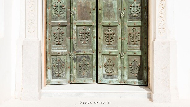 Uma porta dupla com detalhada ferragem metálica e motivo de cruz, emoldurada por uma estrutura arquitetônica clássica.