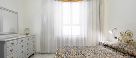 Un dormitorio con cama, cómoda, mesita de noche, lámpara y ventana con cortinas.