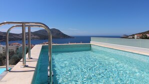 En swimmingpool med en metallstege, kristallklart blått vatten och en utsikt över havet och kullarna i bakgrunden.