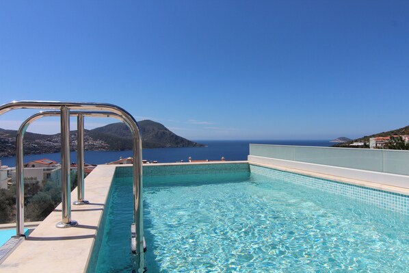 A swimming pool with a metal ladder, clear blue water, and a view of the sea and hills in the background.