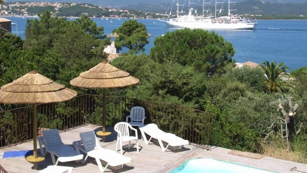 Un espace piscine doté de chaises longues et de parasols en chaume, offrant une vue sur une étendue d’eau où l’on aperçoit un voilier et, au loin, des montagnes.