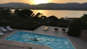 Une piscine où une personne nage, entourée de chaises longues et de parasols, avec une vue sur un coucher de soleil se reflétant sur une étendue d’eau et des montagnes en arrière-plan.