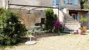 En rustik bygning i sten med en patio, der har et bord og stole, omgivet af grøn vegetation og plantekrukker.