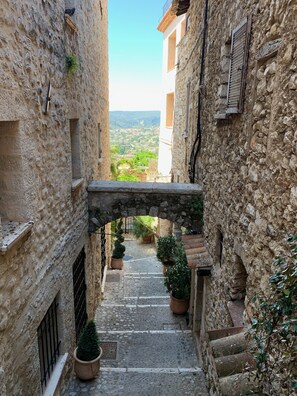 Une étroite ruelle pavée, surmontée d’une arche en pierre rustique, agrémentée de plantes en pot et offrant une vue sur une ville lointaine.