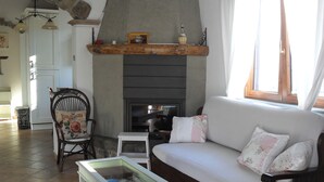 Ein gemütliches Wohnzimmer mit Kamin aus Stein, einem weißen Sofa mit floralen Kissen, einem Holzstuhl und einem Couchtisch mit Glasplatte.