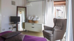 Ein Schlafzimmer mit Bett, Sessel, Kommode, Fernseher und einem Fenster mit Vorhängen.