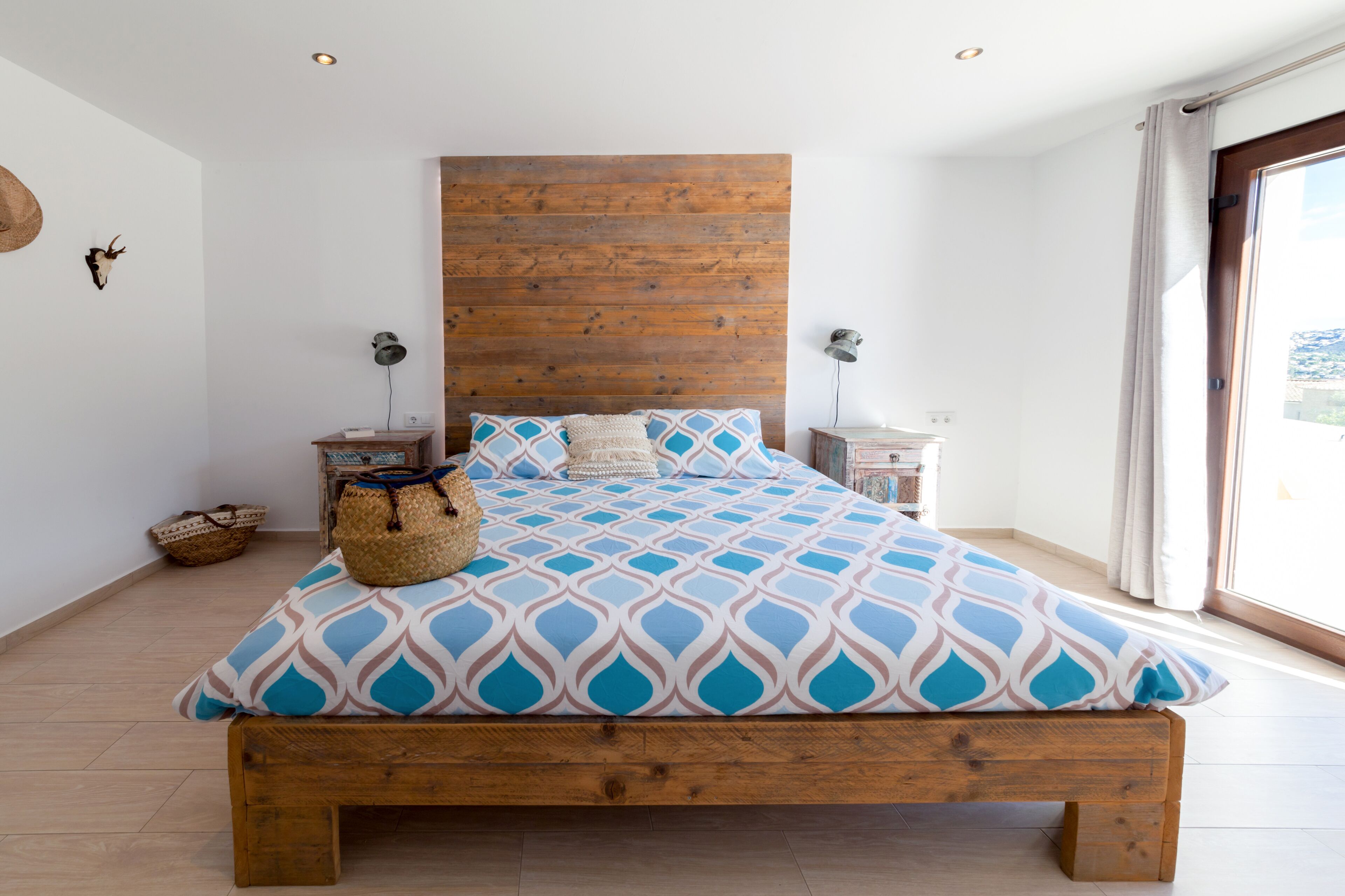 A bedroom with a wooden headboard, a patterned bedspread, and a view through a large window.